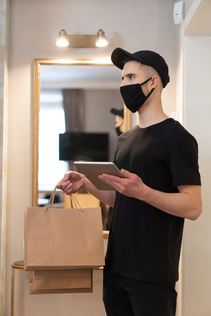 A delivery man with a mask holding paper bags and a tablet, standing indoors.