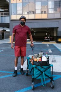 Delivery person with grocery cart in urban setting, wearing mask for pandemic safety.